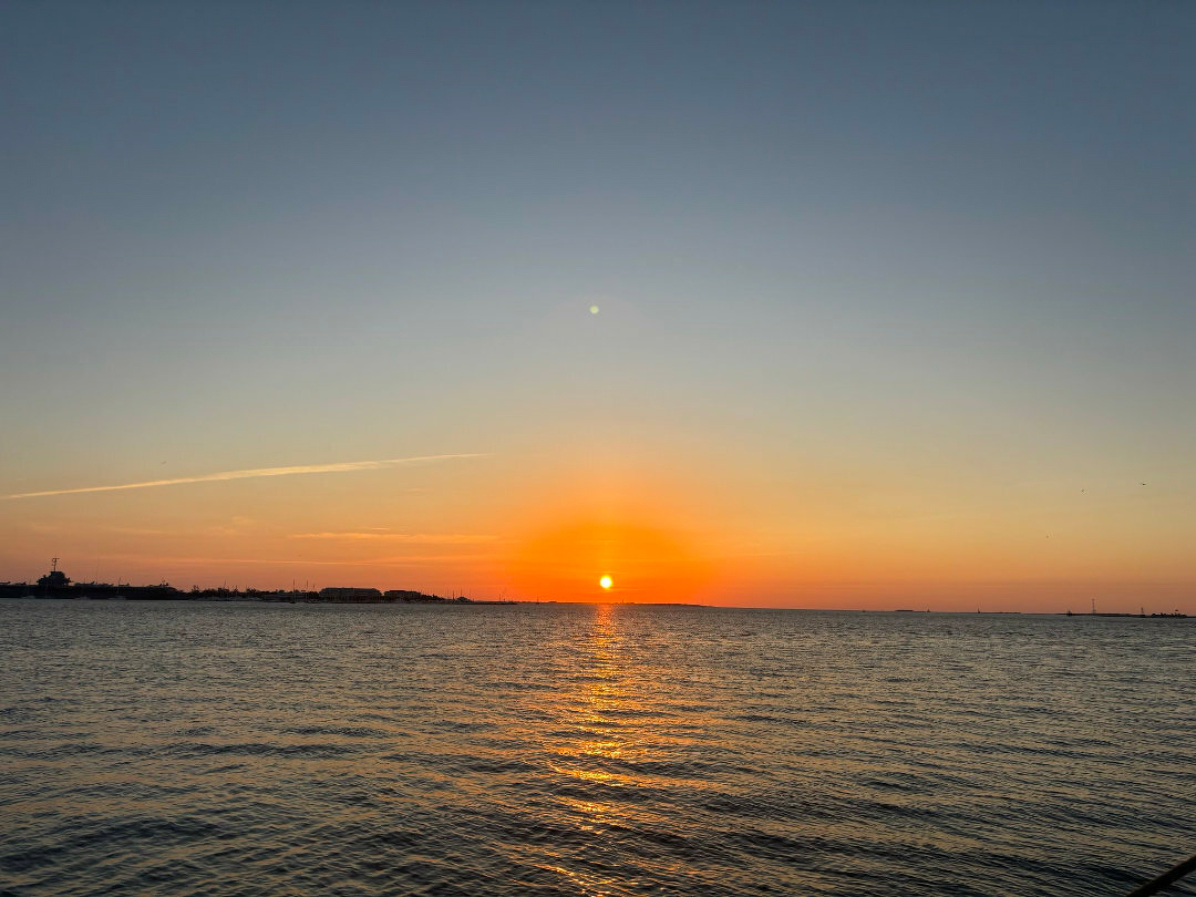 Auto-generated description: A serene sunset over a calm body of water with the sun visible just above the horizon.