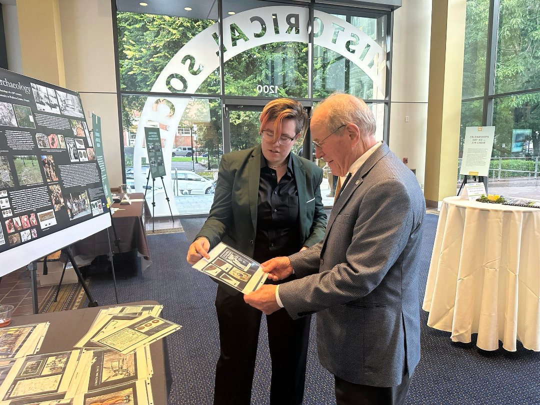 Auto-generated description: Two people are reviewing documents at a display set up inside a building with large windows.