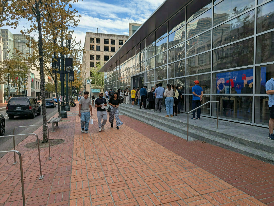 Auto-generated description: People are waiting in line outside a large glass-fronted building (the Apple store) on a city street.