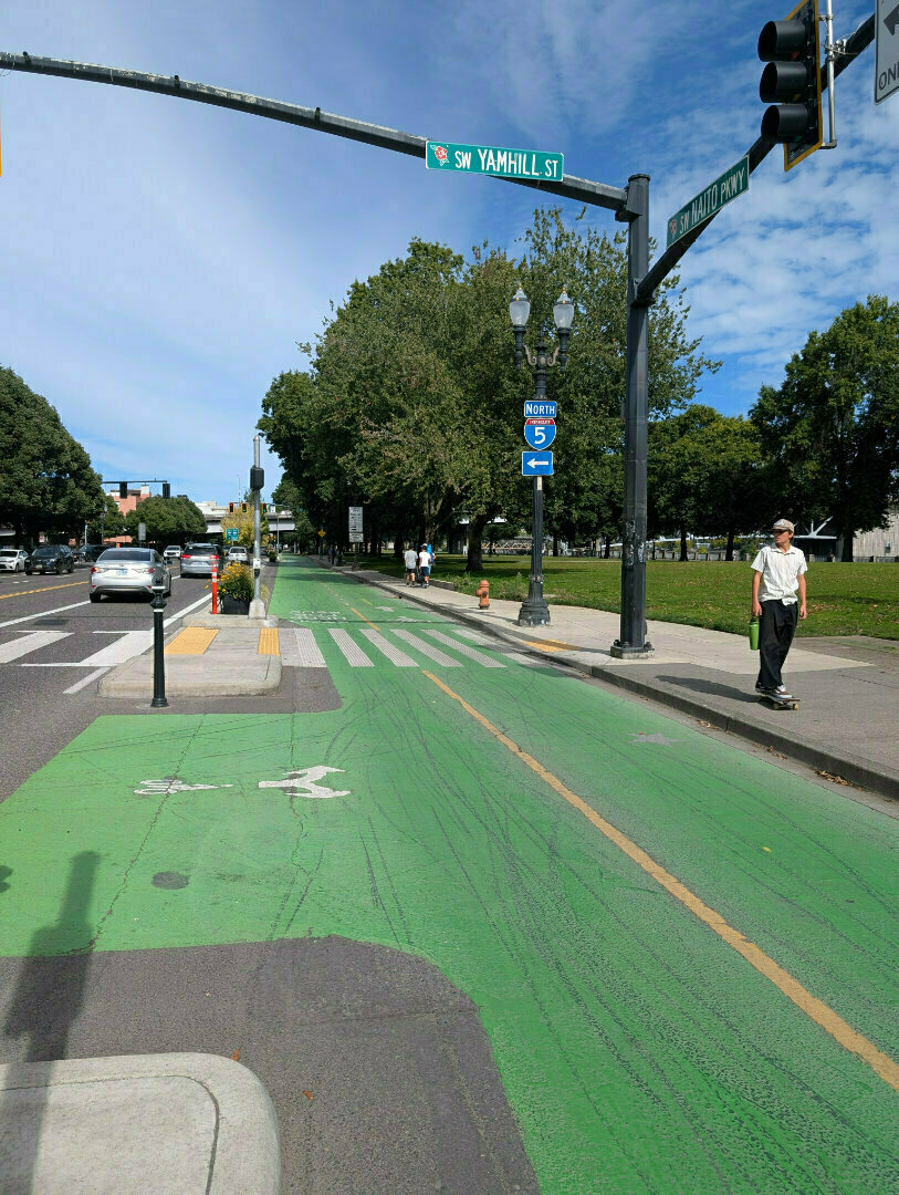 Auto-generated description: A green bike lane runs alongside a street intersection with pedestrian and traffic lights, as people walk on the sidewalk and cars wait at the stoplight.