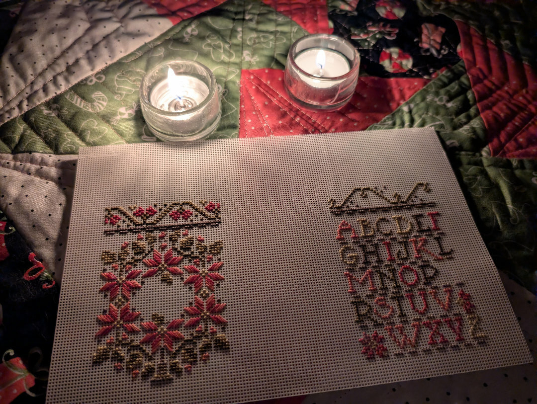 Auto-generated description: A piece of cross-stitch embroidery featuring decorative patterns and the alphabet is set on a quilted surface with two lit candles nearby.