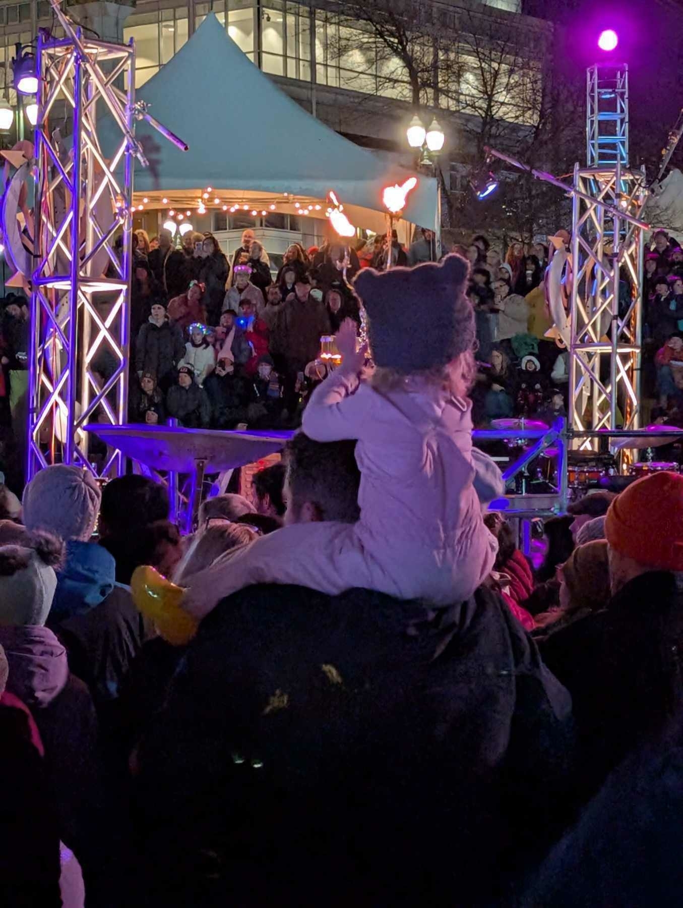 Auto-generated description: A child wearing a winter hat sits on someone's shoulders, watching a performance with flame jugglers at an outdoor event.