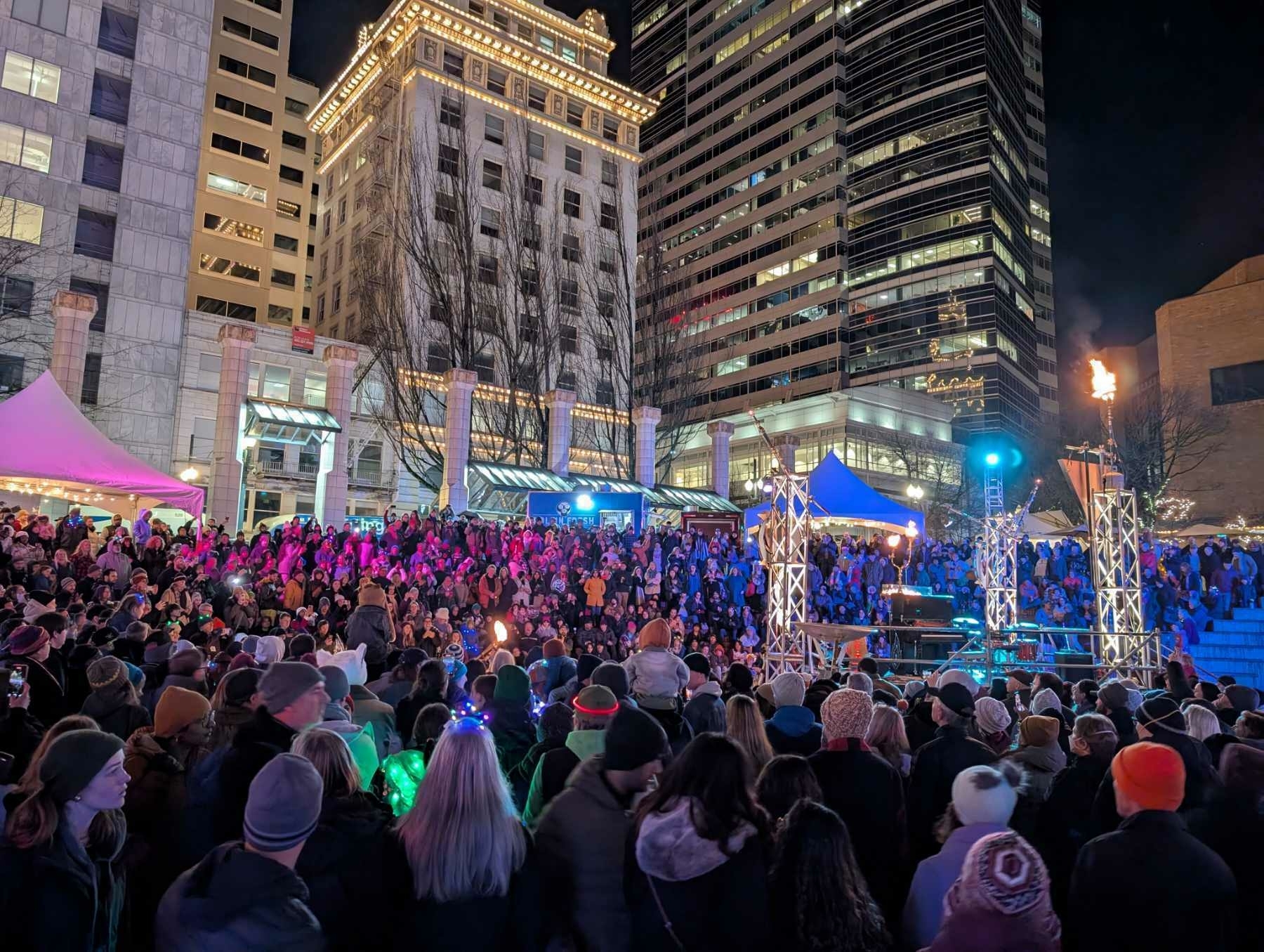 Auto-generated description: A large crowd gathers in a brightly lit urban square at night, surrounded by tall buildings and festive lighting.