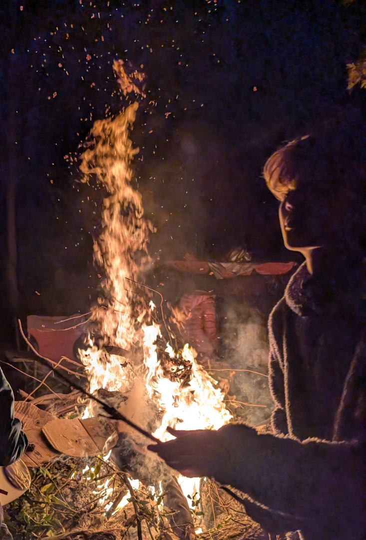 Auto-generated description: A person is standing by a campfire at night, holding a stick, with the flames and sparks illuminating the scene.