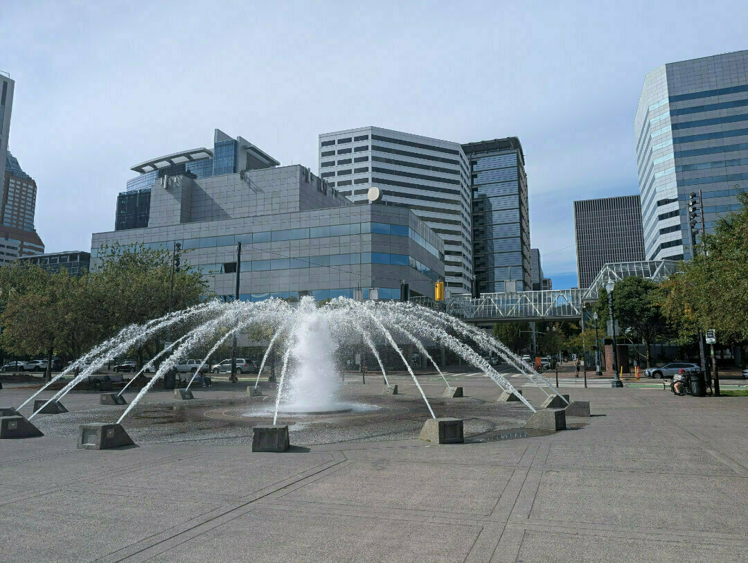 Auto-generated description: A geometric fountain shoots water upward in a plaza surrounded by modern, glass-fronted buildings.