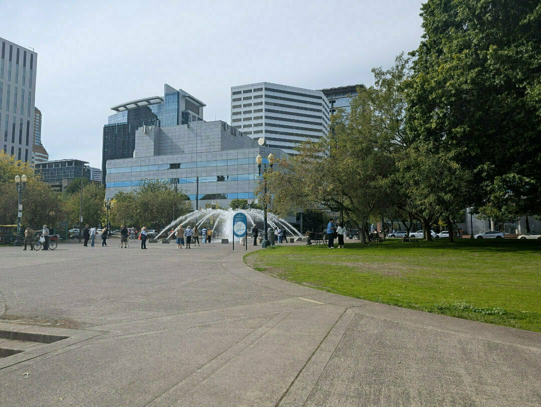 Auto-generated description: A park scene featuring a modern building backdrop, a fountain surrounded by people, and trees bordering a green lawn.