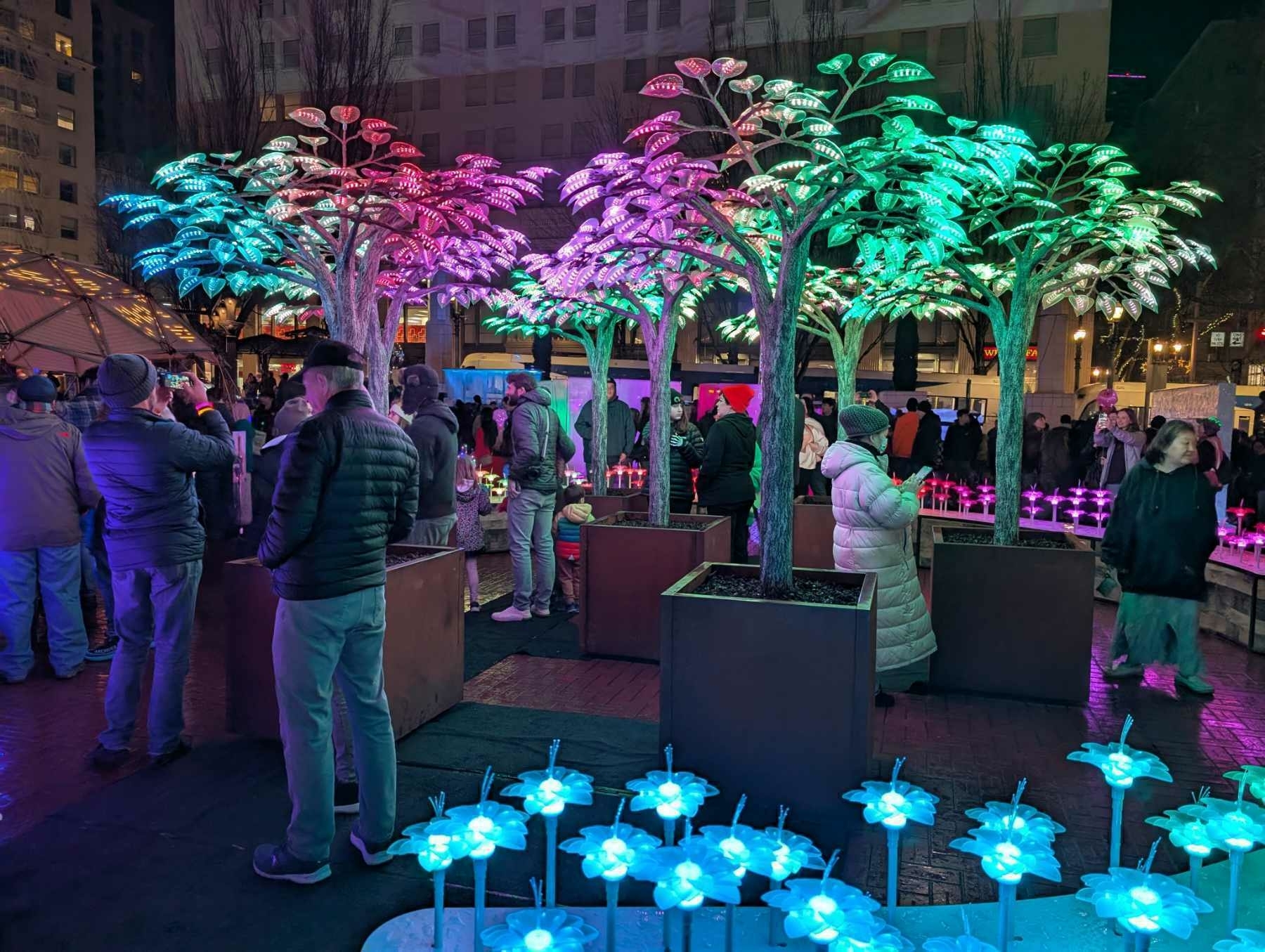 Auto-generated description: People are gathered around brightly lit, colorful artificial trees at a night event.