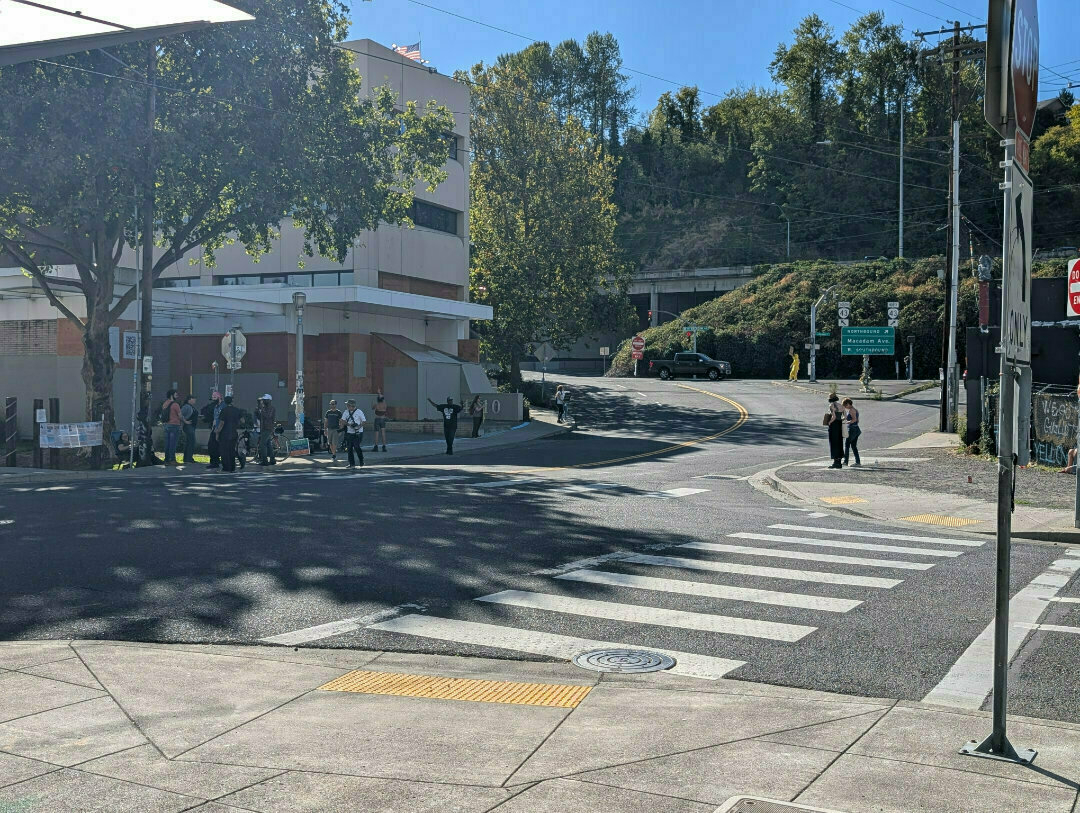 Auto-generated description: A group of people are gathered near a crosswalk at a sunny intersection with trees and buildings in the background.