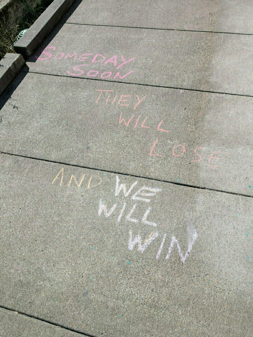 Auto-generated description: Colorful chalk text on a sidewalk reads, SOMEDAY SOON THEY WILL LOSE AND WE WILL WIN.