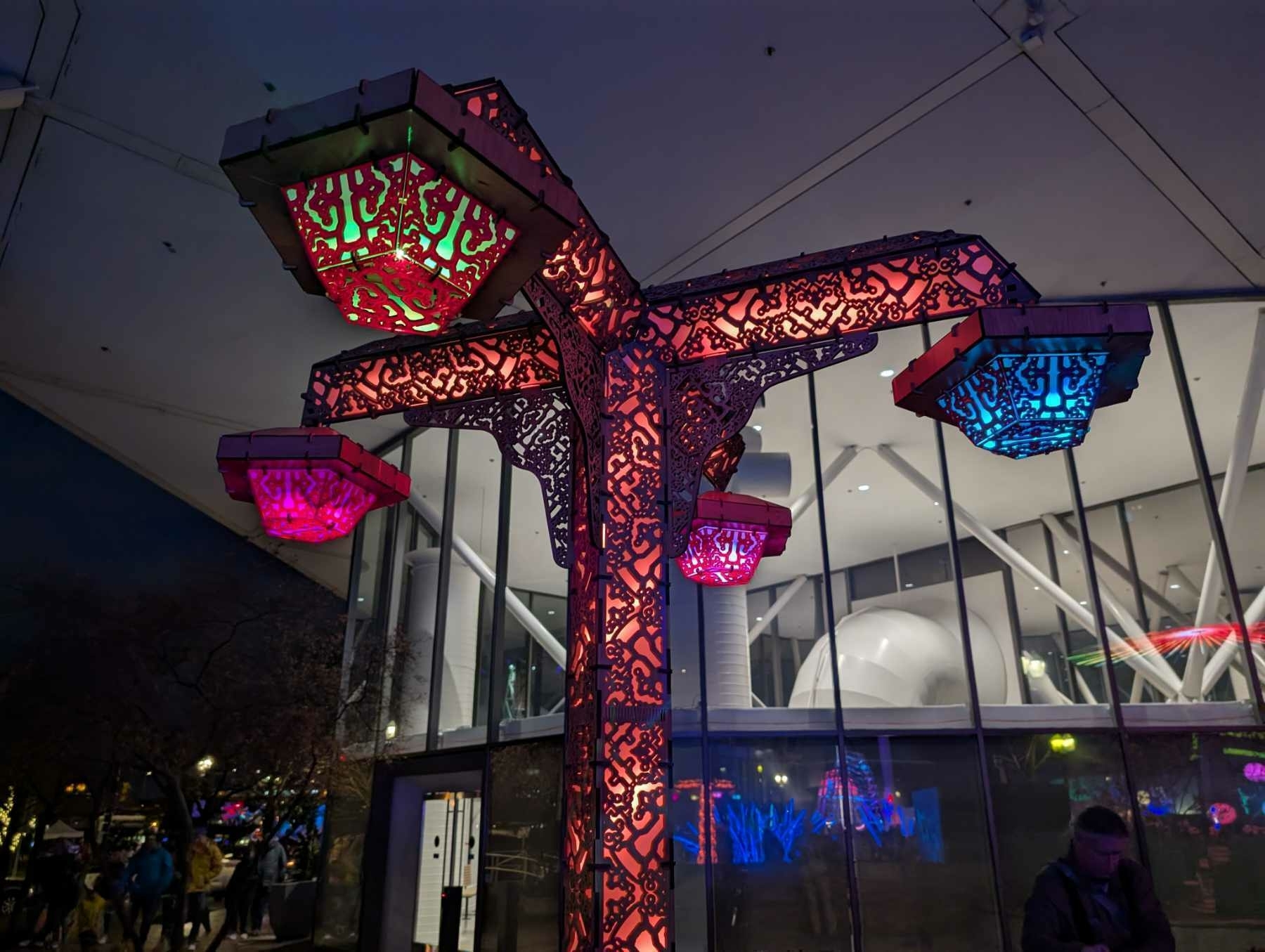 Auto-generated description: A decorative, illuminated structure with colorful lights stands near a modern glass building at dusk.