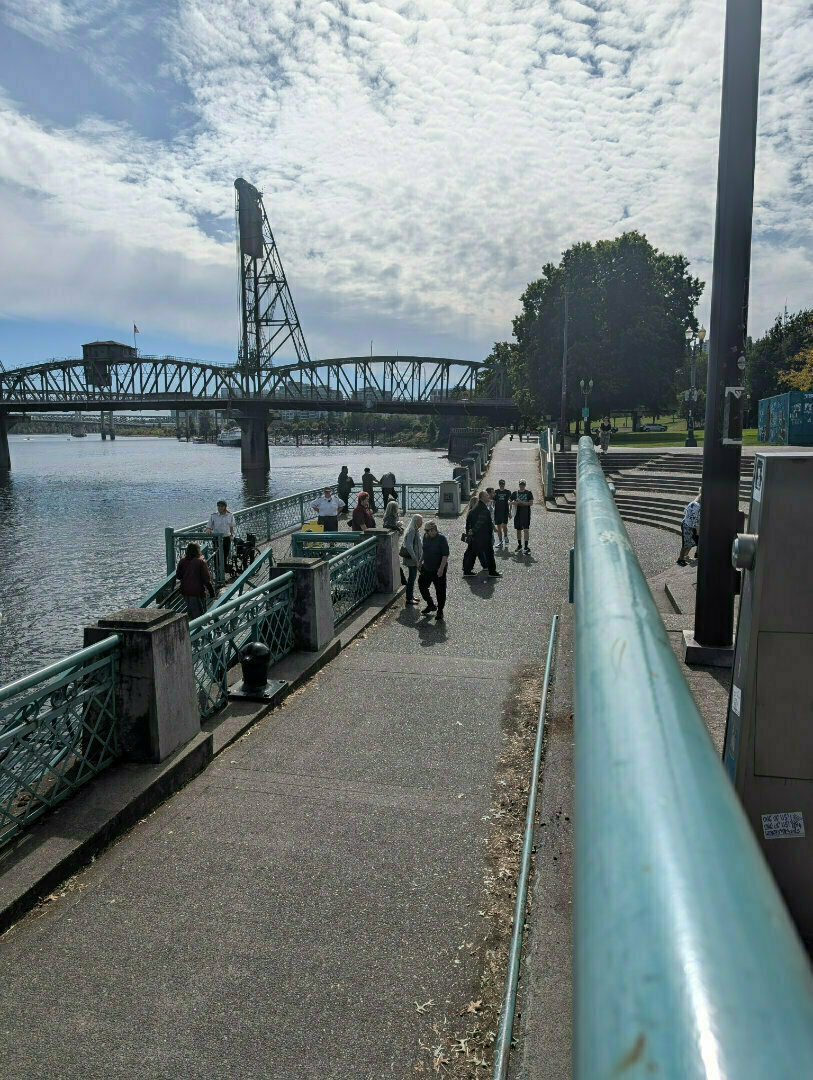 Auto-generated description: People are walking along a riverside pathway near a bridge on a cloudy day.