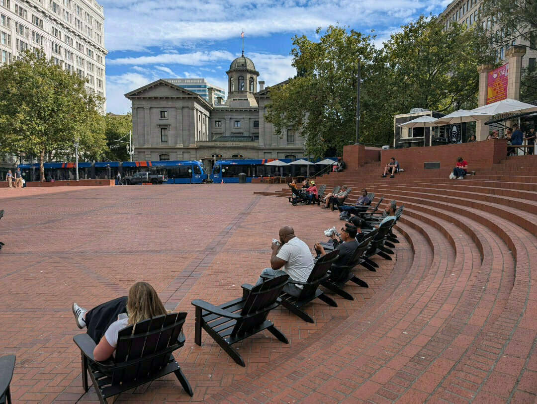 Auto-generated description: People are relaxing on chairs in a large brick-paved open square surrounded by historic buildings and trees.