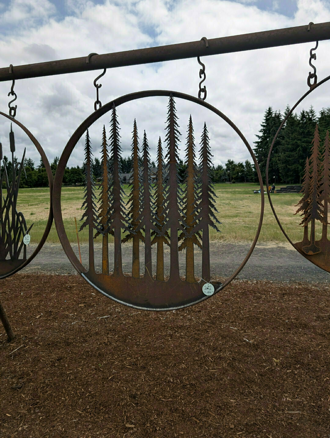 Auto-generated description: A metal circular art piece featuring silhouettes of tall pine trees is hanging outdoors.