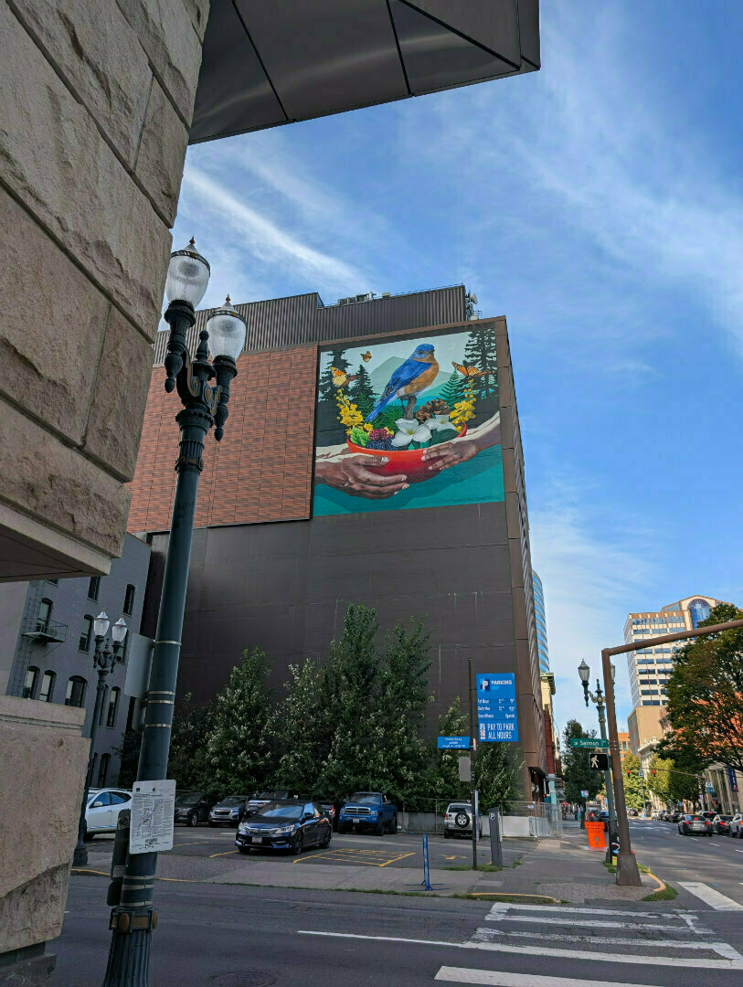 Auto-generated description: A vibrant mural featuring a bird perched on a hand, surrounded by plants, adorns the side of a building in an urban setting.