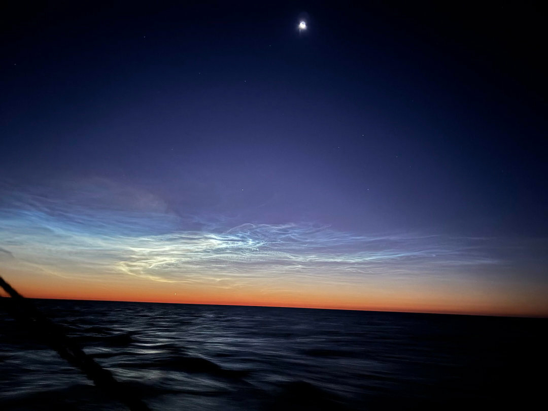 Auto-generated description: A twilight ocean scene features faint noctilucent clouds glowing against a deep blue sky, with a small bright celestial object above.