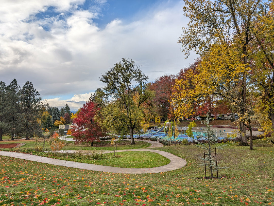 Auto-generated description: A winding path leads through a park with vibrant autumn foliage and a playground under a partly cloudy sky.