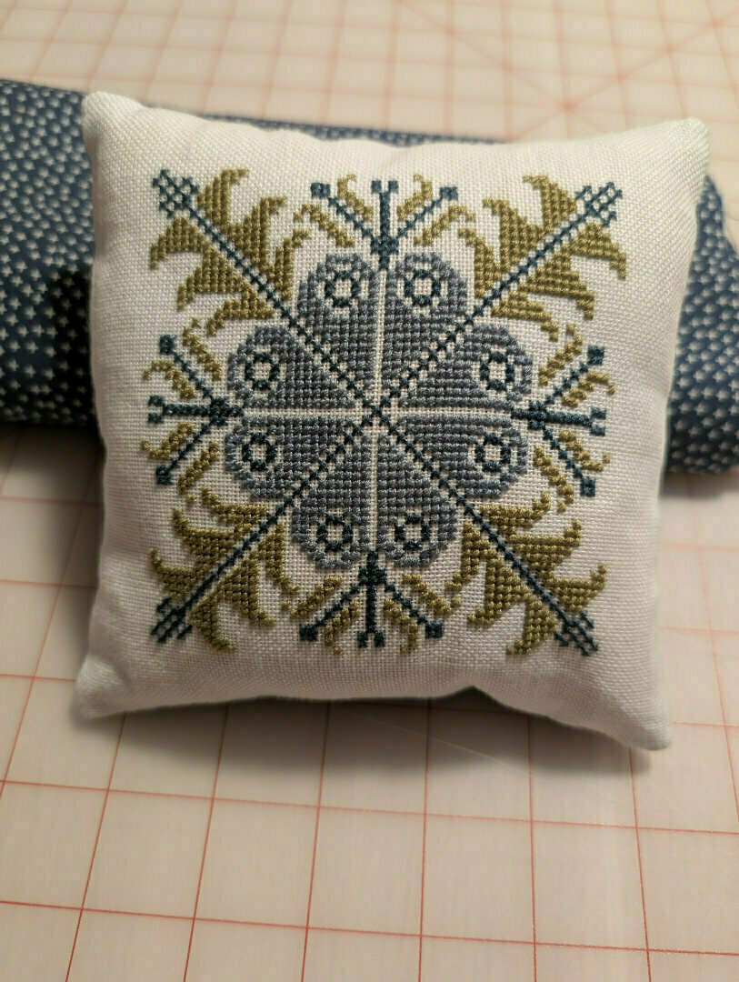 Auto-generated description: A small pillow features an intricate cross-stitched pattern in blue and green, resting on a patterned fabric.