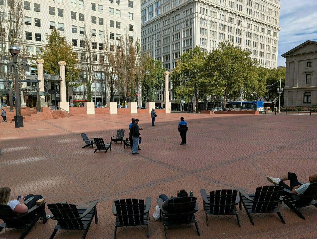 Auto-generated description: People are seated on chairs in a spacious brick-paved square surrounded by tall buildings and trees.