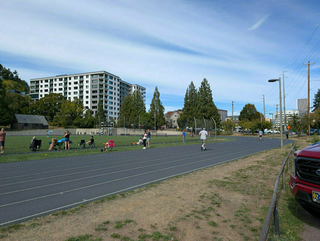 Auto-generated description: People are exercising and relaxing near a track in an urban park with buildings and trees in the background.