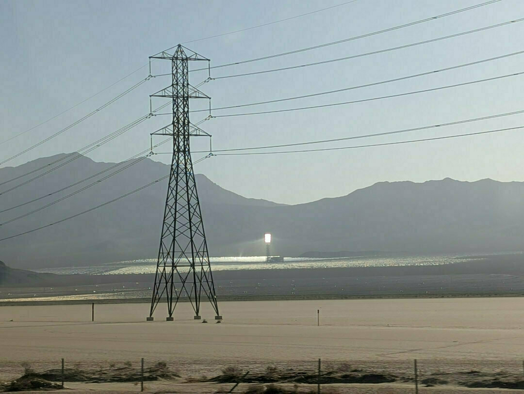 Auto-generated description: A solar power tower is seen in the distance, surrounded by reflective panels in a desert landscape with mountains and a transmission tower.