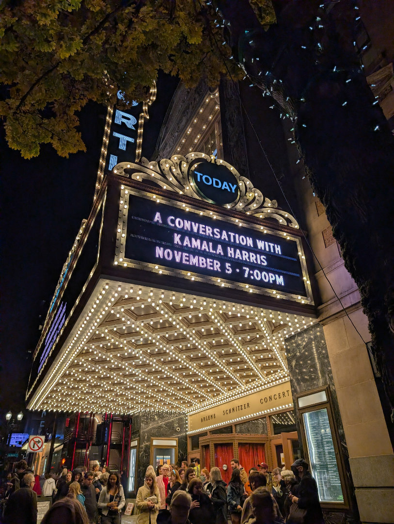 Auto-generated description: A theater marquee brightly lit at night announces A Conversation with Kamala Harris on November 5th at 7:00 PM, with a crowd gathering below.