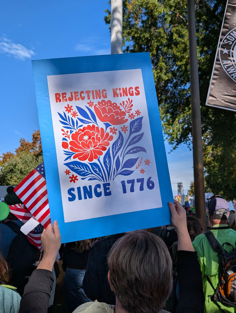 Auto-generated description: A person is holding a colorful protest sign with floral designs, reading Rejecting Kings Since 1776 at an outdoor gathering.