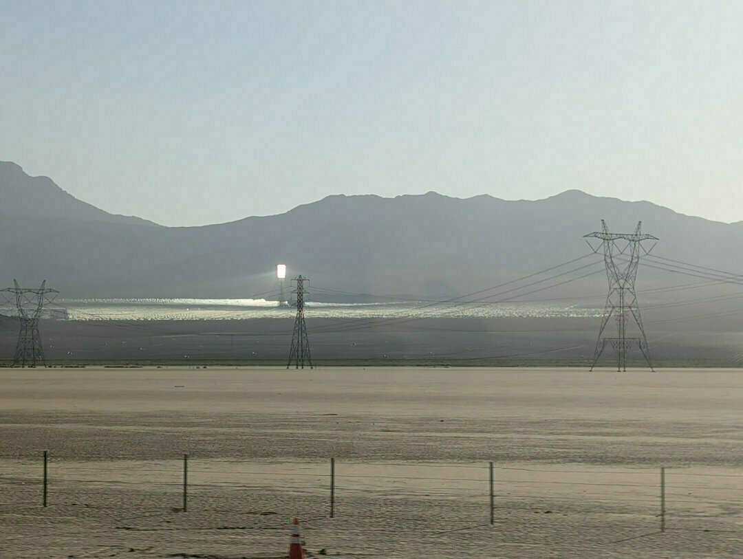 Auto-generated description: A solar power plant is situated in a desert landscape, with transmission towers and power lines visible against distant mountains.