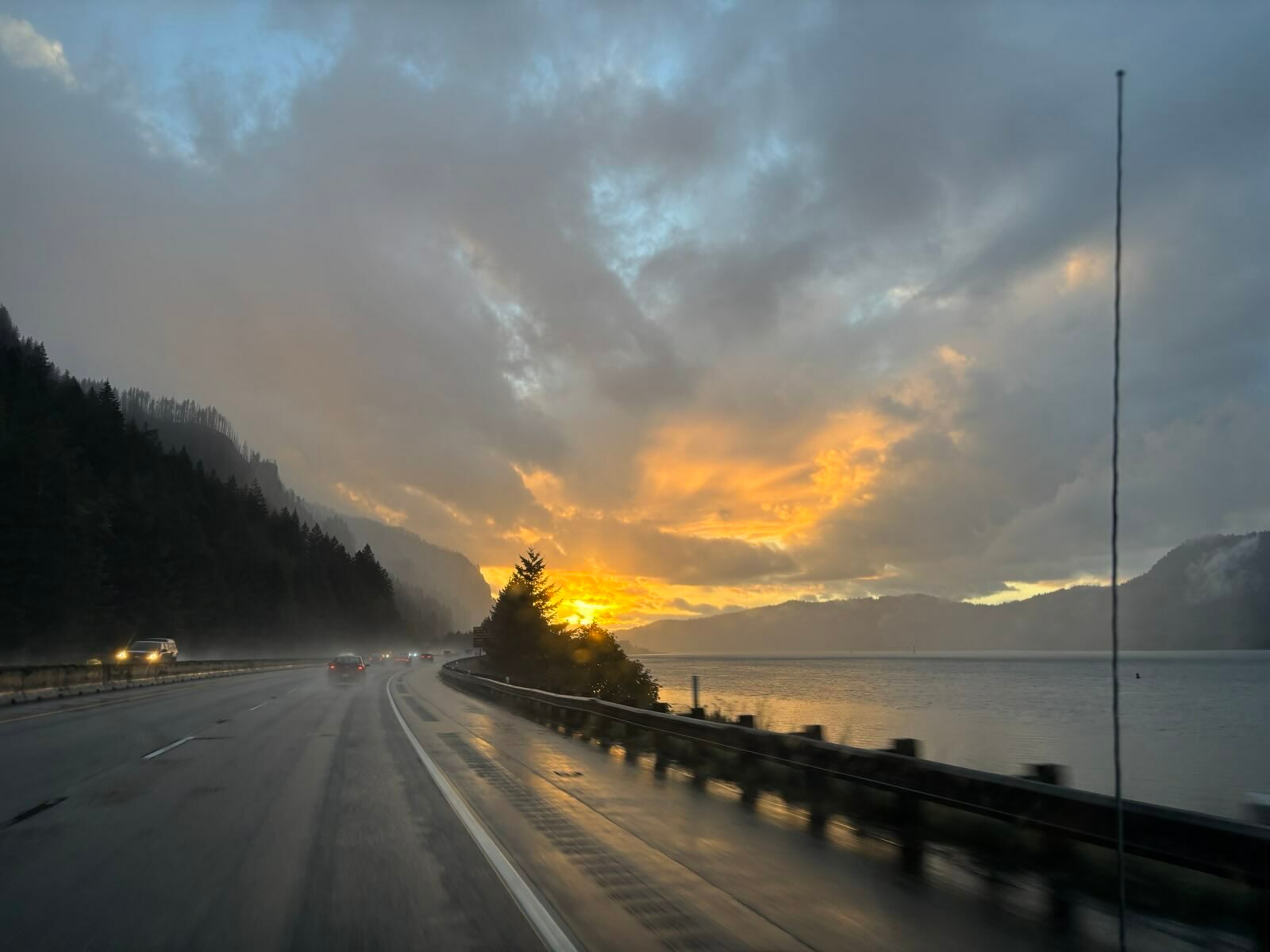 Auto-generated description: A scenic highway runs alongside a lake with a dramatic sunset breaking through the clouds.