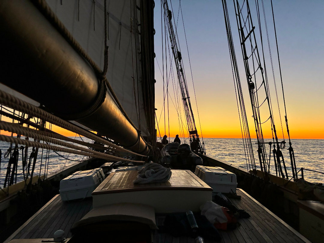 Auto-generated description: A sailing ship deck is silhouetted against a vibrant sunset over the ocean.