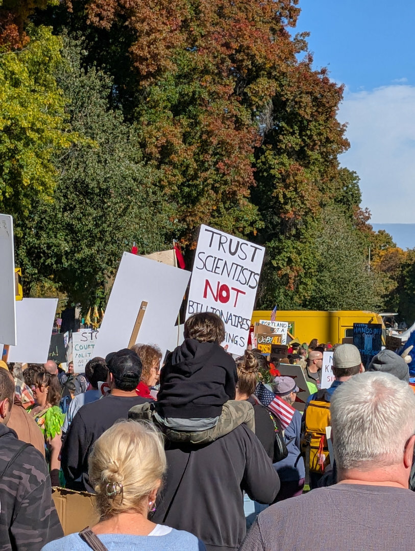 Auto-generated description: A crowd gathers at a protest with a variety of signs, one prominently referencing scientists and billionaires.