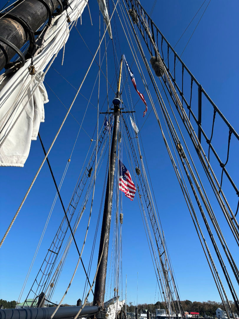 Auto-generated description: Tall ship rigging and masts with an American flag waving against a clear blue sky.