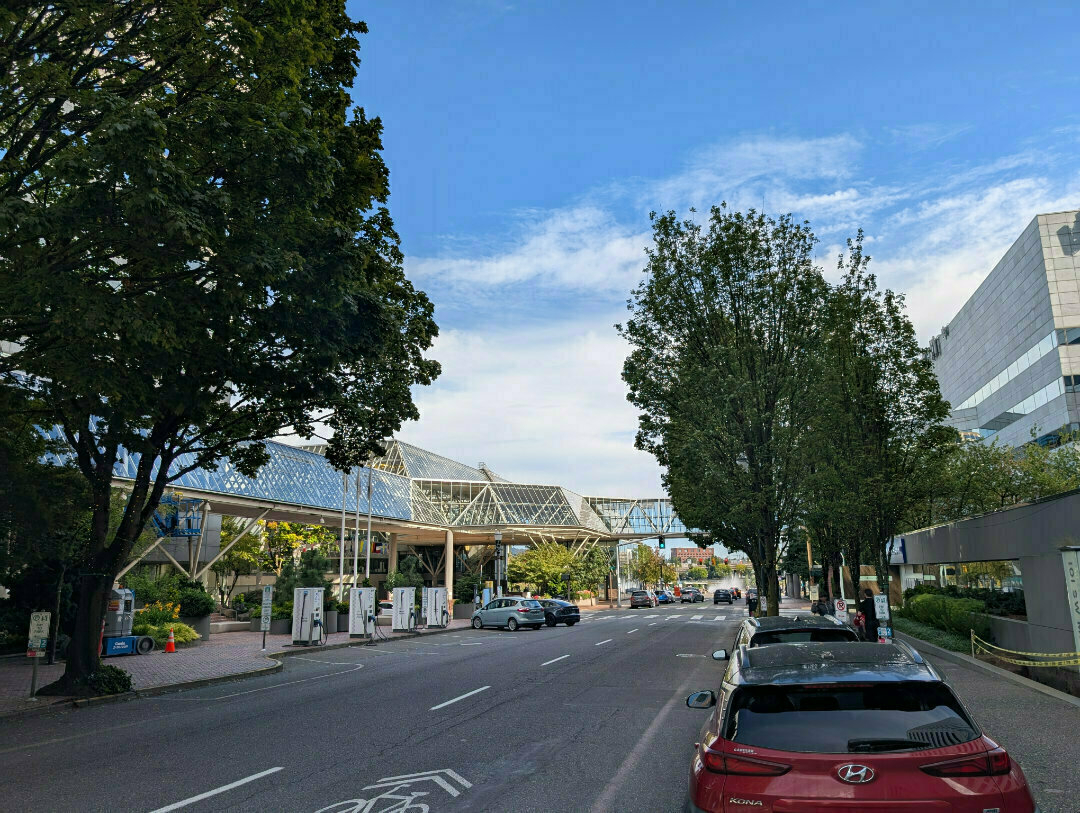 Auto-generated description: A tree-lined urban street features parked cars, a modern glass pedestrian bridge, and pedestrians walking nearby.