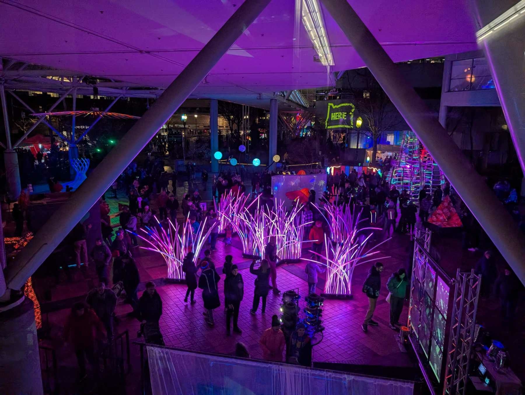 Auto-generated description: People gather in a vibrant outdoor space with colorful purple and blue lighting, featuring art installations and illuminated decorations.