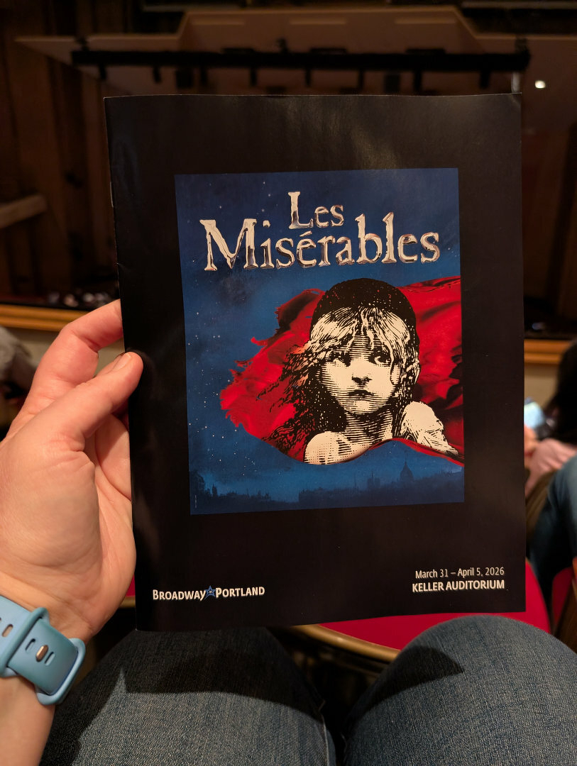 Auto-generated description: A person is holding a program for Les Misérables at the Keller Auditorium, which is scheduled for performances from March 31 to April 5, 2026.