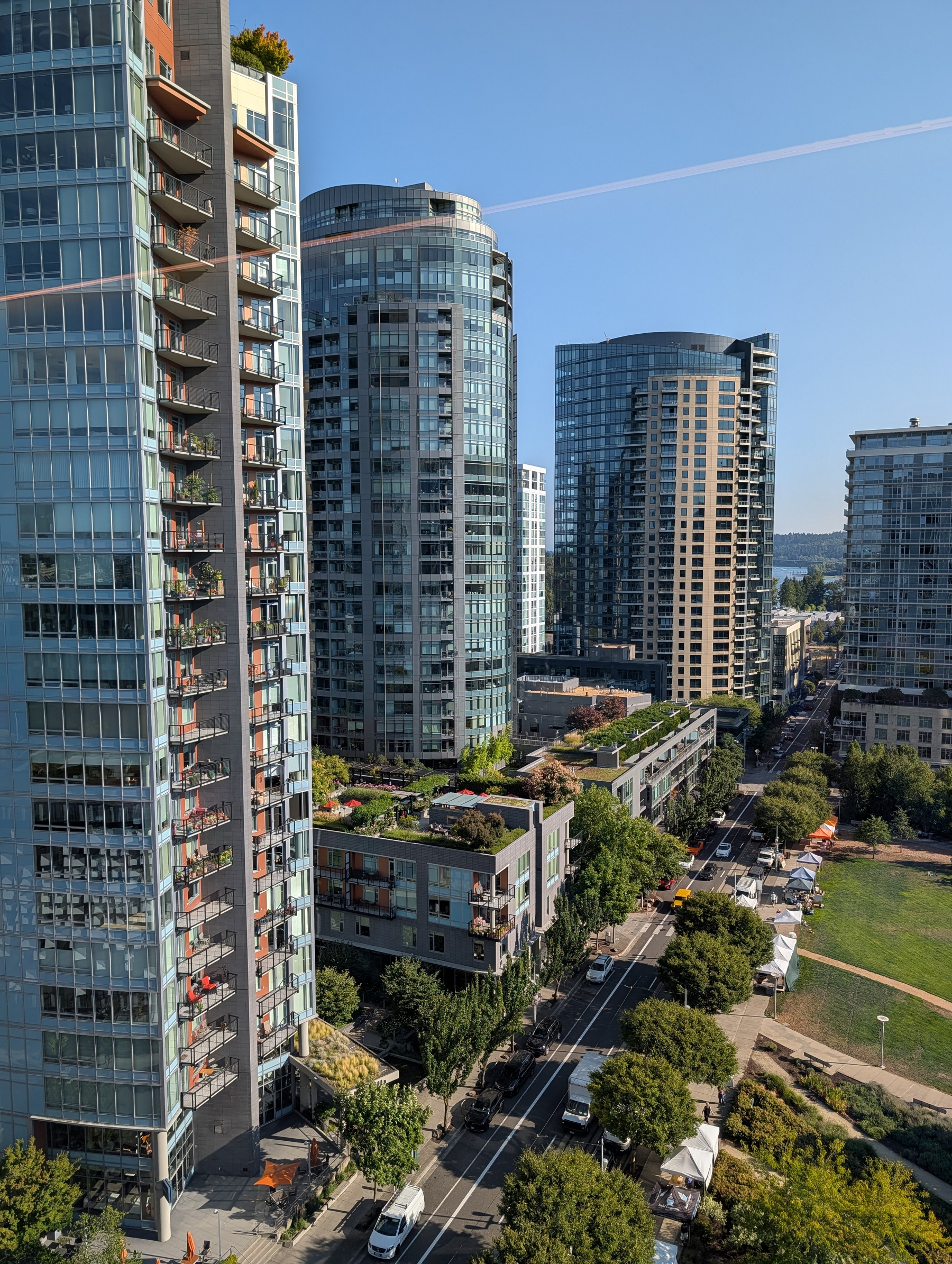 A cityscape features modern high-rise buildings with balconies, a tree-lined street with vehicles, and a green park area under a clear blue sky.