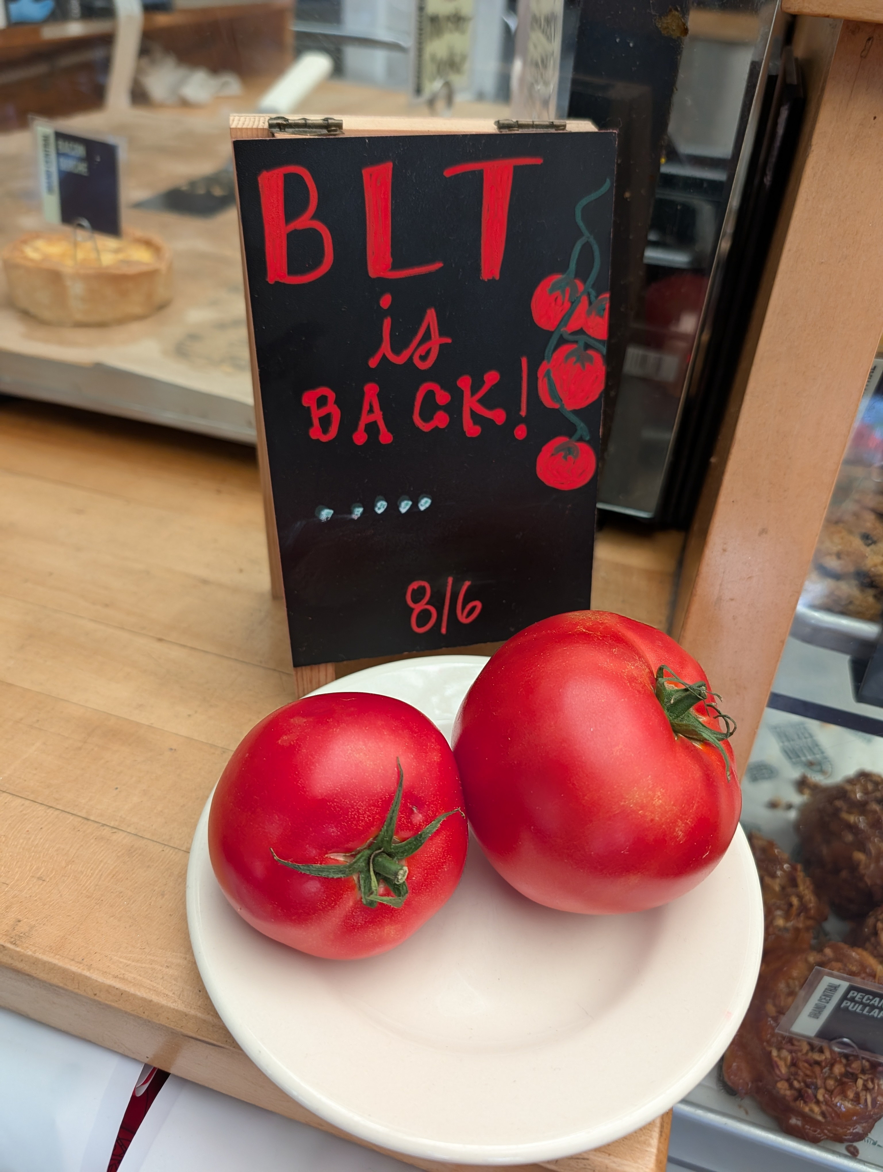 Two ripe tomatoes are placed on a white plate in front of a sign that announces BLT is BACK! with a drawing of tomatoes.