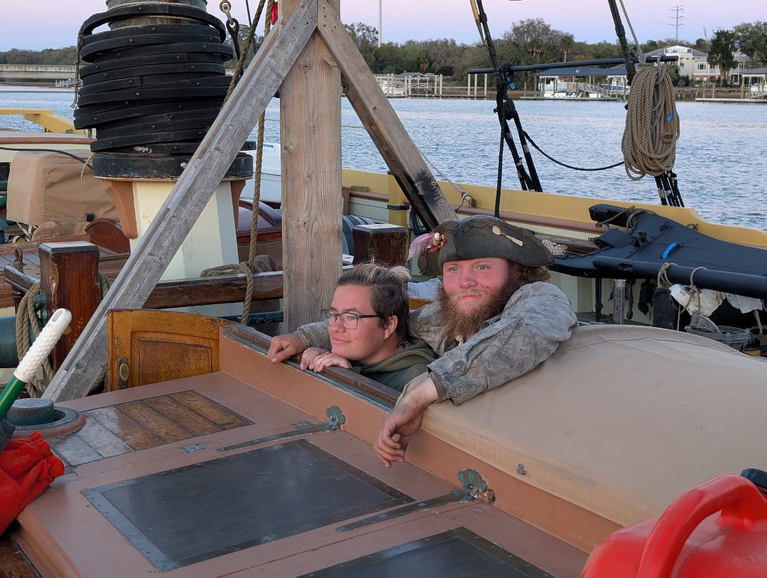 Auto-generated description: Two people are peeking out from a wooden structure on a boat, with water and other boats in the background.