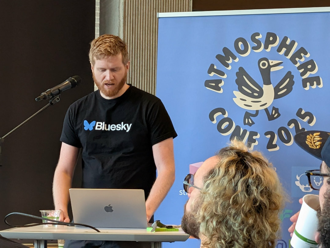 Auto-generated description: A person is speaking at a podium with a Atmosphere Conf 2025 banner in the background.