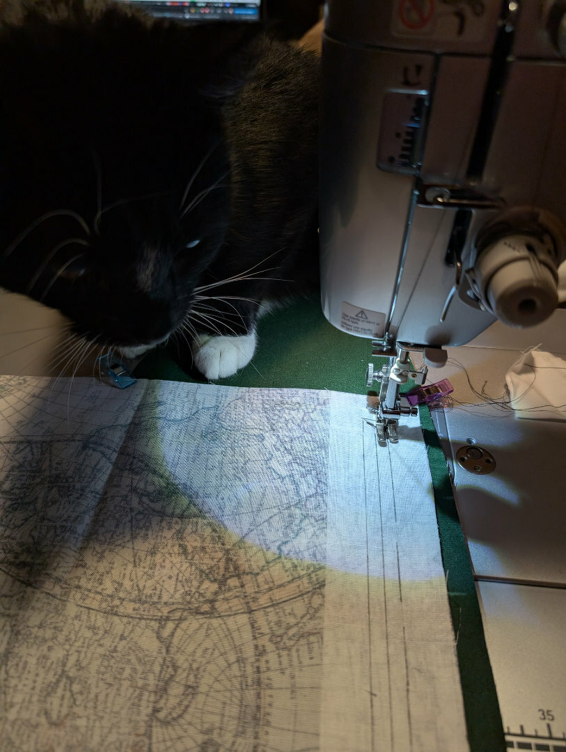 Auto-generated description: A black and white cat curiously observes a sewing machine working on fabric featuring a map design.
