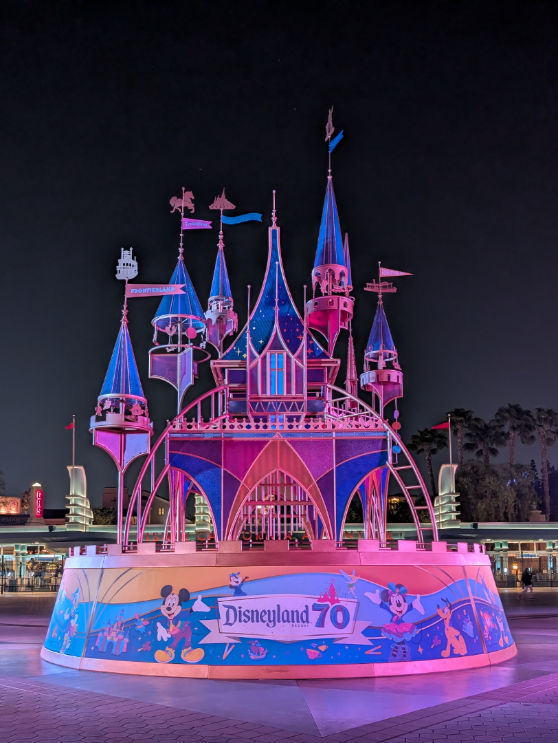 Auto-generated description: A whimsical castle decoration adorned with colorful lights and Disneyland 70 signage is set against a nighttime backdrop.
