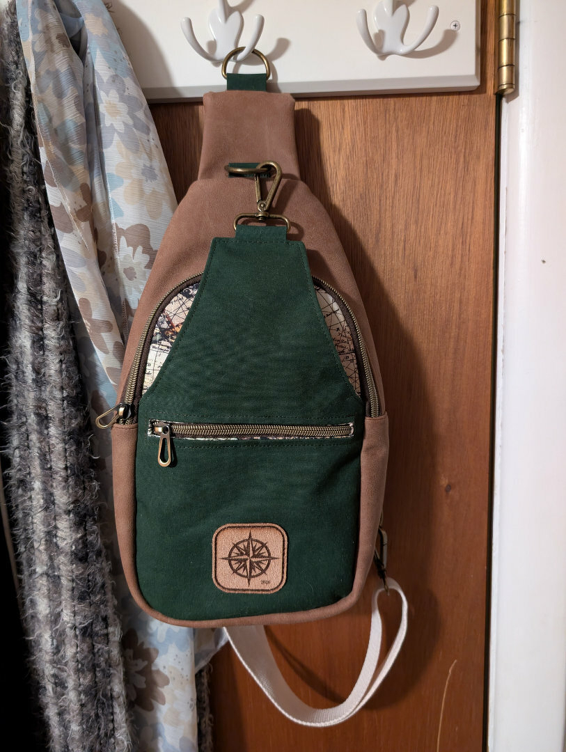 Auto-generated description: A stylish sling bag with green and brown colors hangs on a wooden door, featuring a front zipper pocket and a compass design patch.