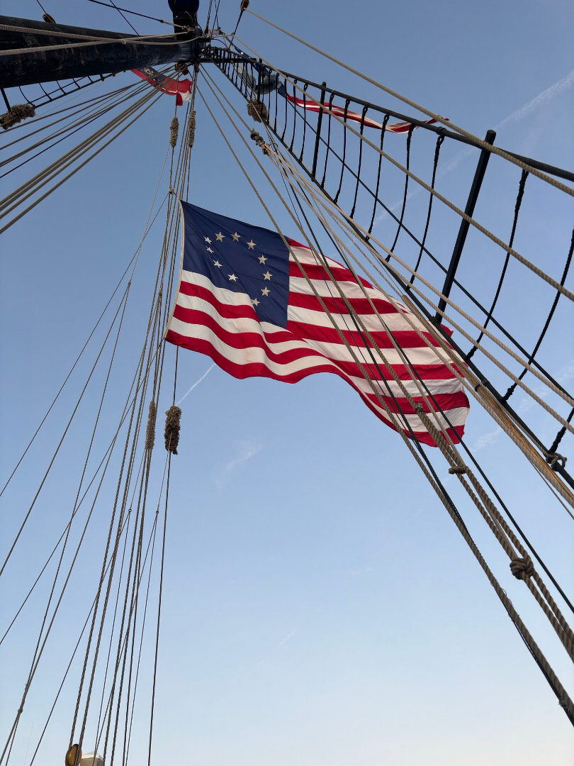 Auto-generated description: A historic American flag with thirteen stars and stripes is hoisted on a ship's rigging against a clear blue sky.