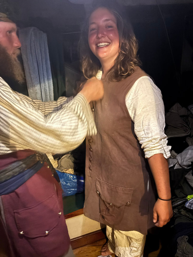 Auto-generated description: A person is smiling while another person helps adjust their vest-like garment in a dimly lit room.