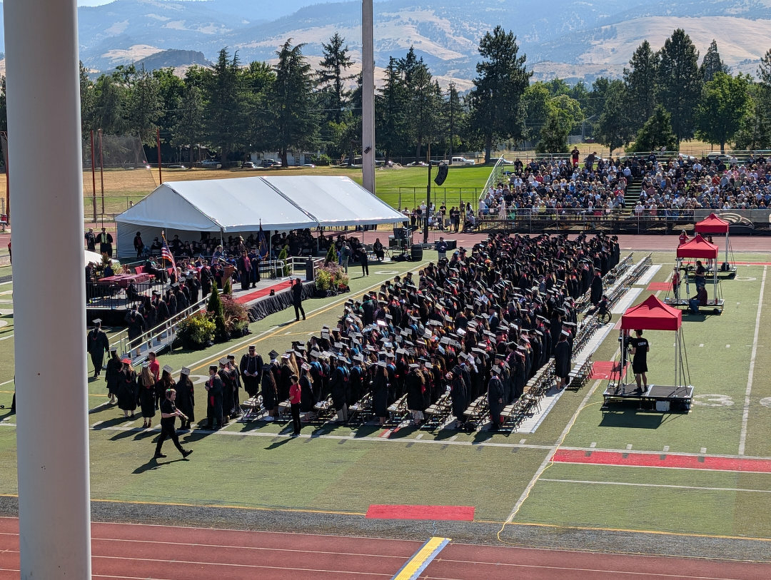 Auto-generated description: A graduation ceremony is taking place on a sports field, featuring graduates in caps and gowns, an audience in bleachers, and white canopies over podiums.
