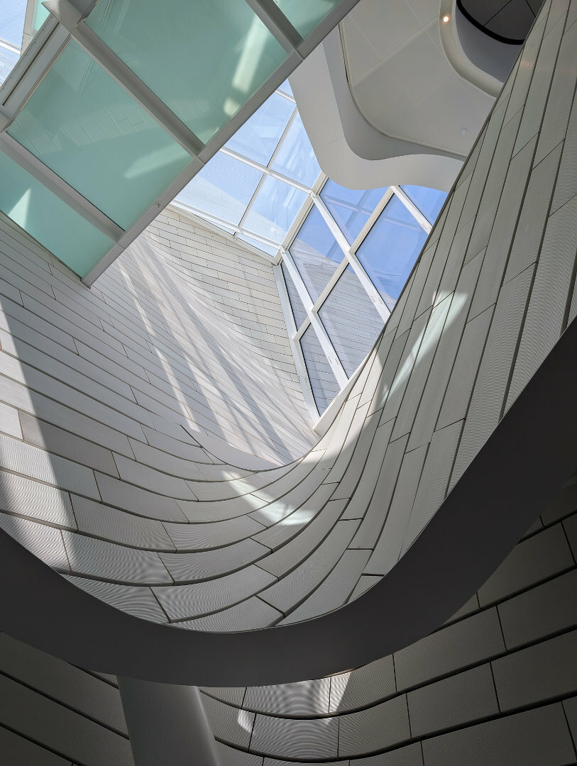 Auto-generated description: A modern architectural interior showcases an upward view of a curving, textured wall and large glass windows, allowing natural light to stream in.