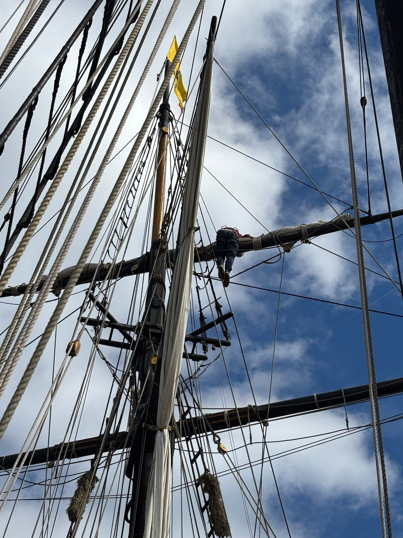 Auto-generated description: A person is climbing the rigging of a tall ship against a backdrop of a partly cloudy sky.