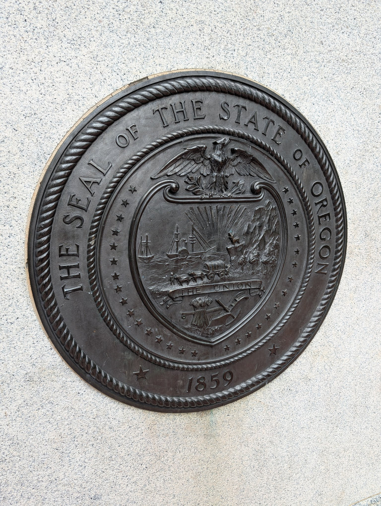 Auto-generated description: A bronze seal of the State of Oregon, dated 1859, is mounted on a stone wall.