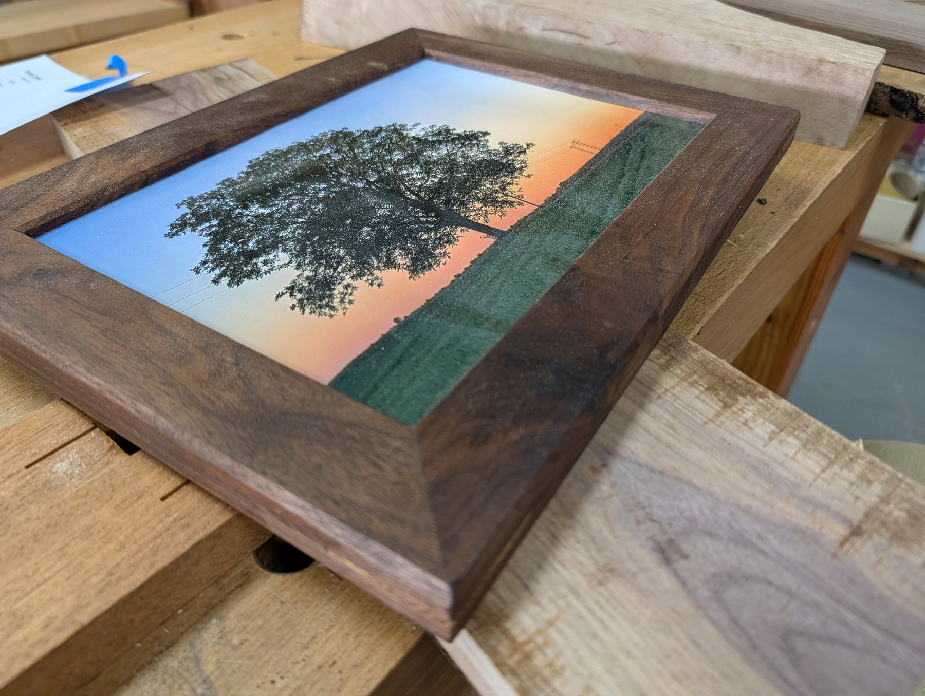Auto-generated description: A framed photograph featuring a tree against a colorful sunset sky is placed on a wooden workbench.
