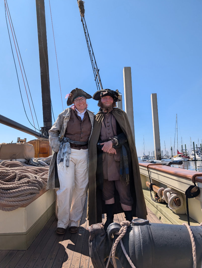 Auto-generated description: Two individuals dressed in period pirate costumes stand on a wooden ship's deck with masts and ropes in the background.