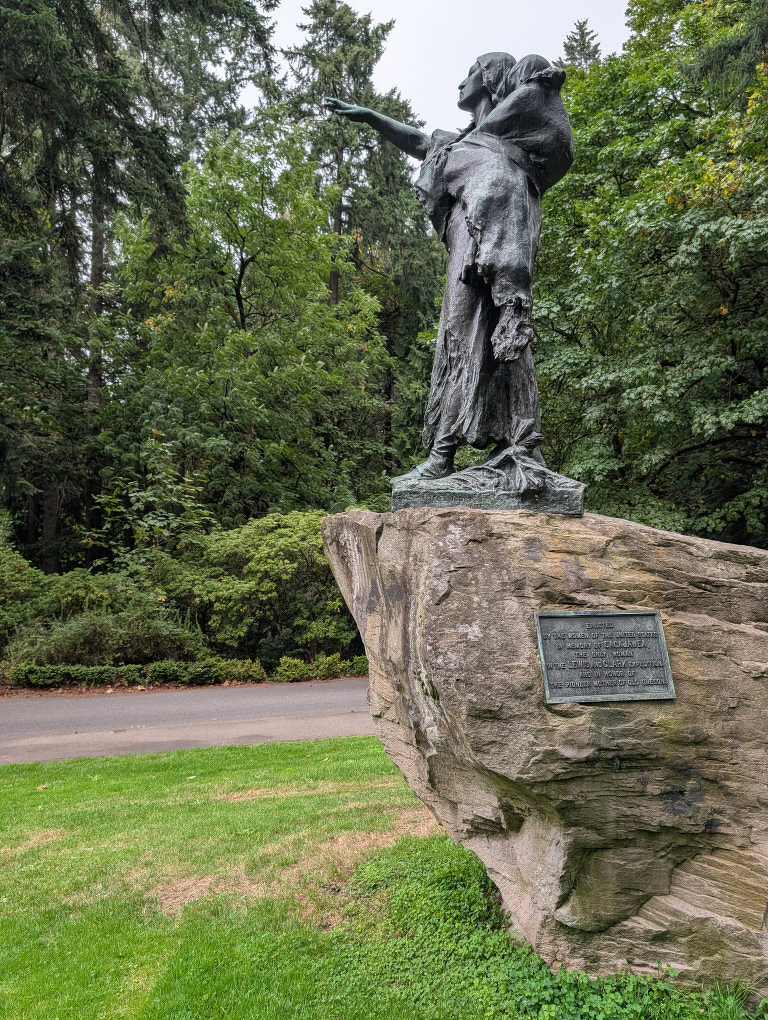 Auto-generated description: A bronze statue of a woman carrying a bundle on her back and reaching out with her arm is mounted on a large rock with a plaque, set in a lush, green park.